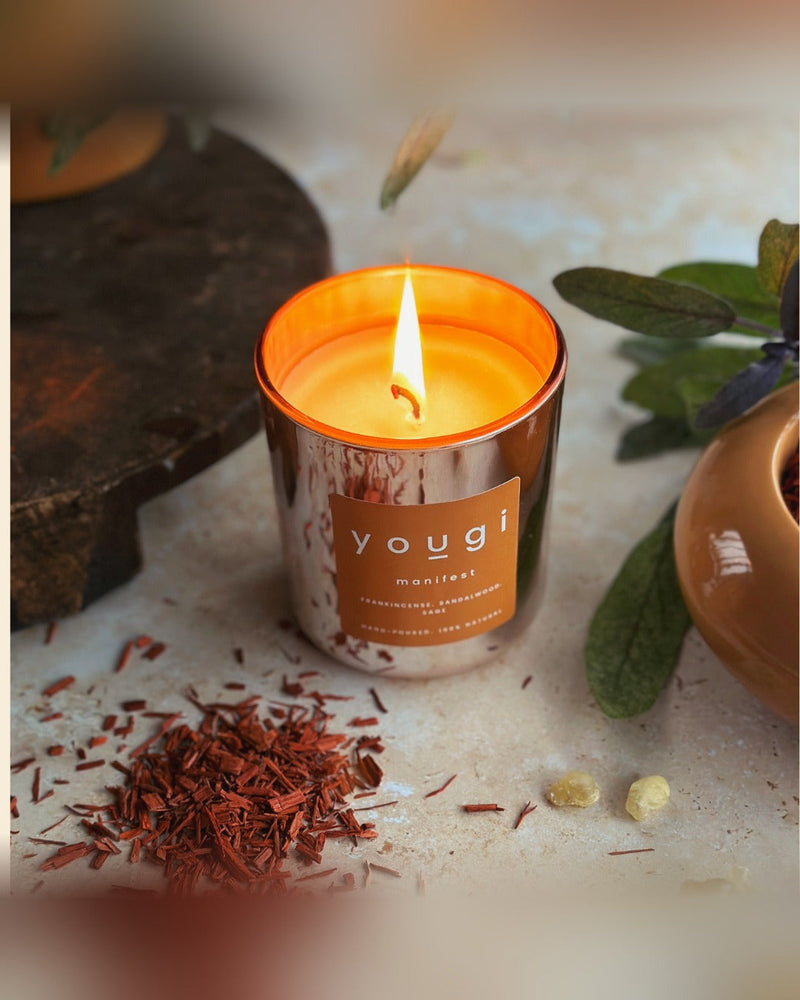 A lit Manifest candle beside sandalwood shavings, the frankincense, sandalwood and sage blend