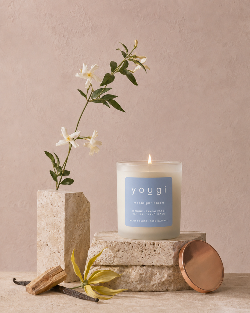A lit Yougi candle in a clear glass jar sits on stacked stone blocks, with a matching round copper lid leaning beside it. White flowers in a stone vase and a vanilla bean add styled props against a soft blush background.