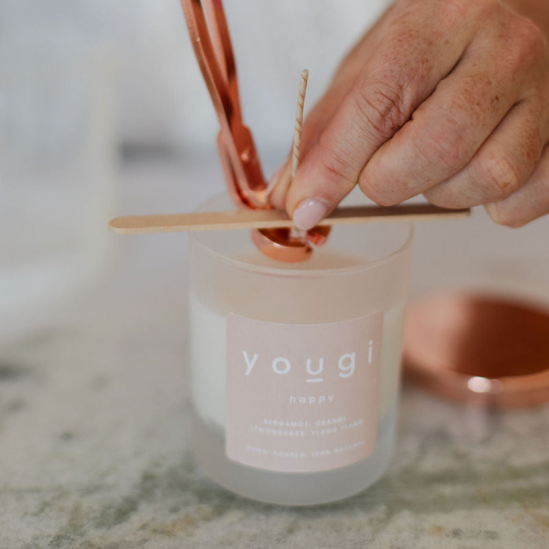 A close-up of hands using a rose gold metal wick trimmer to trim a wick on a frosted glass candle jar with a white label reading 'yougi'.