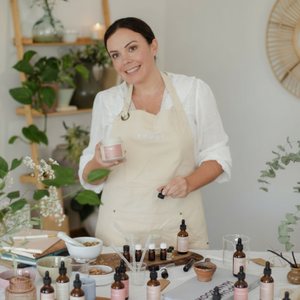 Woman in a white apron holding a product in a room with plants and a clock on the wall.