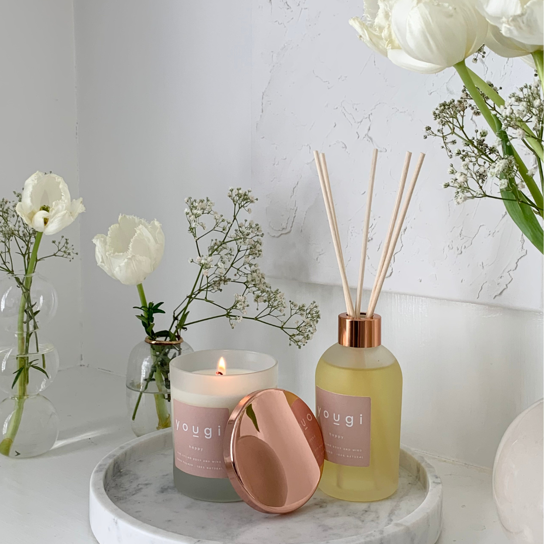 A Yougi reed diffuser with a pink label and a copper cap, alongside a lit candle and white flowers in a vase, on a white marble surface.