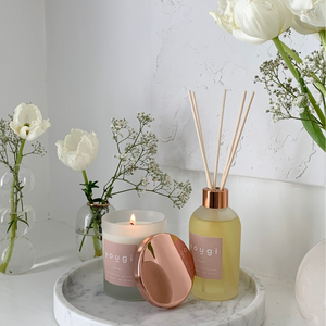 A Yougi reed diffuser with a pink label and a copper cap, alongside a lit candle and white flowers in a vase, on a white marble surface.