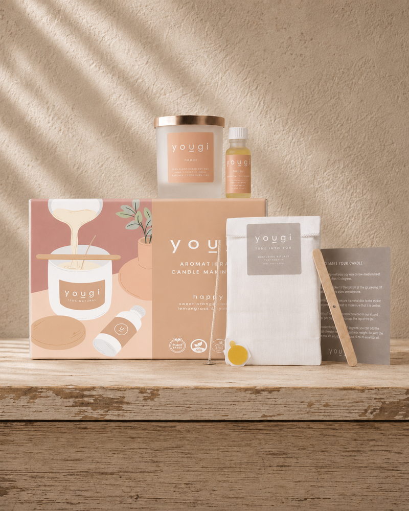 A Yogui Aromatherapy Candle Making kit arranged on a rustic wooden table against a textured beige wall with soft leaf shadows. The kit includes a candle jar with a gold lid, a small oil bottle, a printed box, a white bag of wax, a wooden wick, and a card.