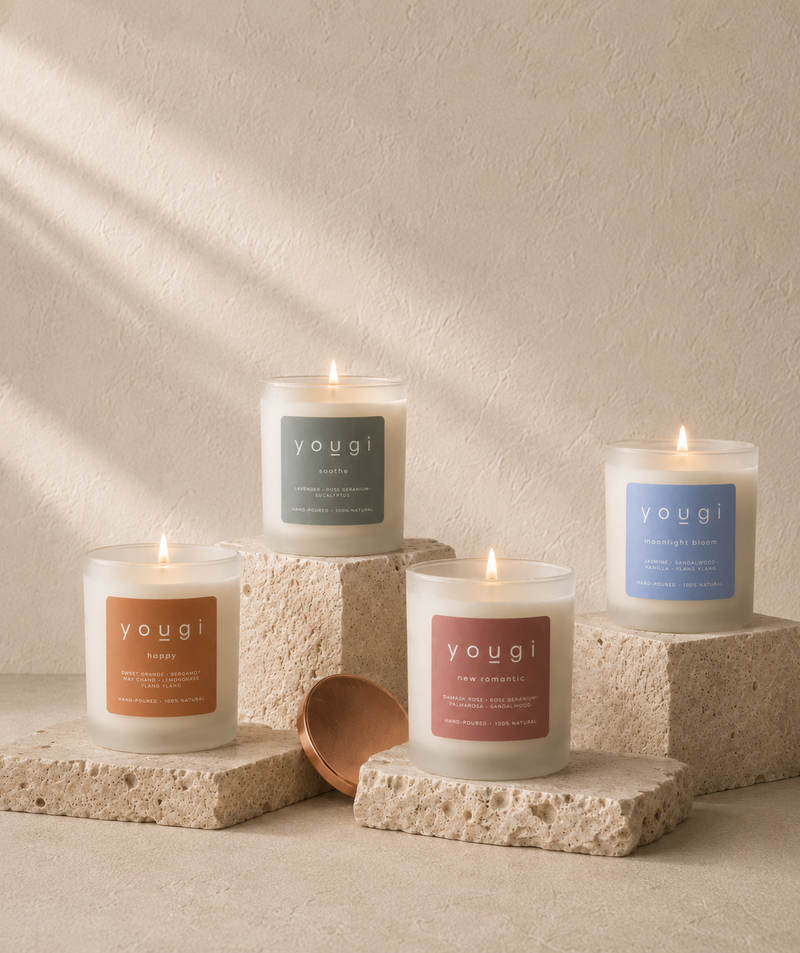 Four Yogui candles in frosted glass jars with lit flames, arranged on textured stone blocks against a warm beige wall with soft shadow patterns. The labels are in muted earthy tones: orange, pink, green, and blue.