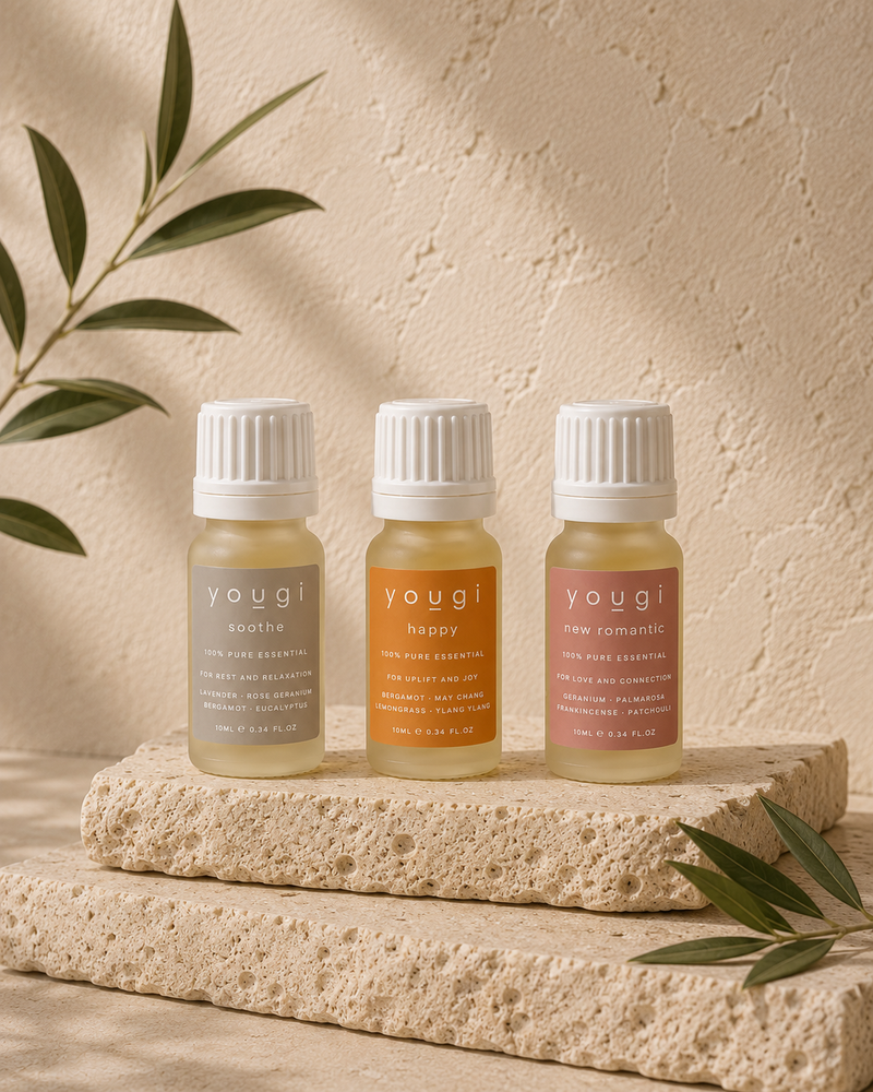 Three small yogui essential oil bottles with white ribbed caps are displayed on a textured stone slab. The bottles contain pale yellow, golden yellow, and dusty rose liquids, with labels reading 'yogui citrus', 'yogui happy', and 'yogui new tomahawk'.