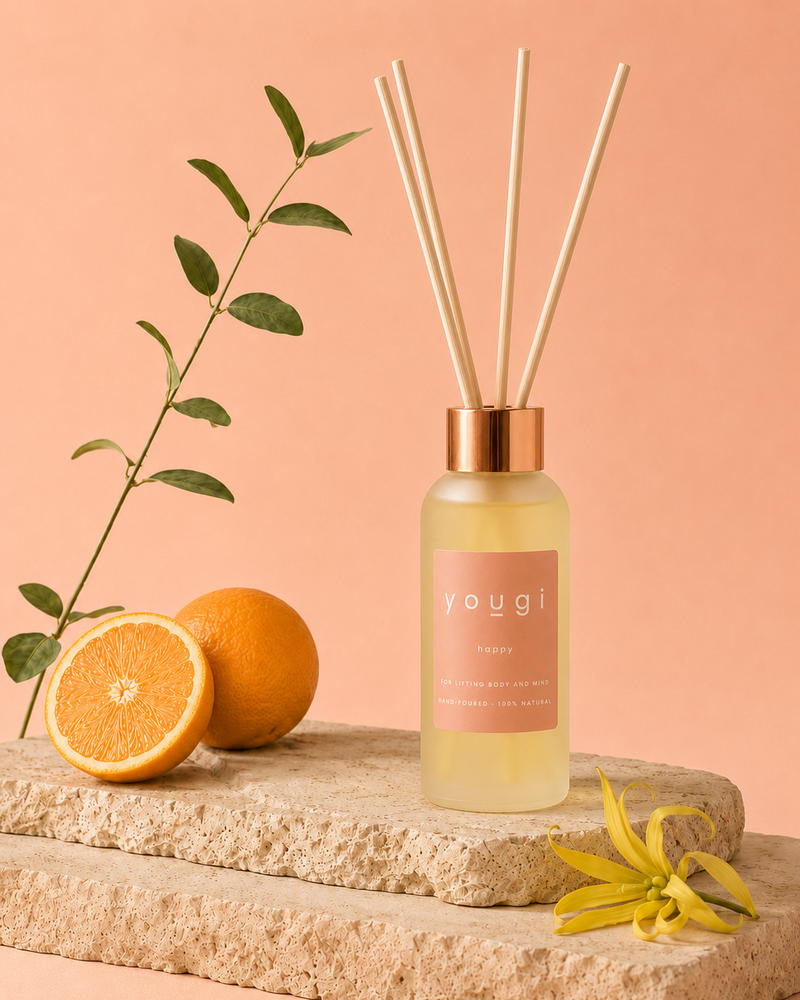A Yougi reed diffuser bottle with a pink label and natural reeds, styled on a stone slab with oranges, a green stem, and a yellow flower against a peach background.