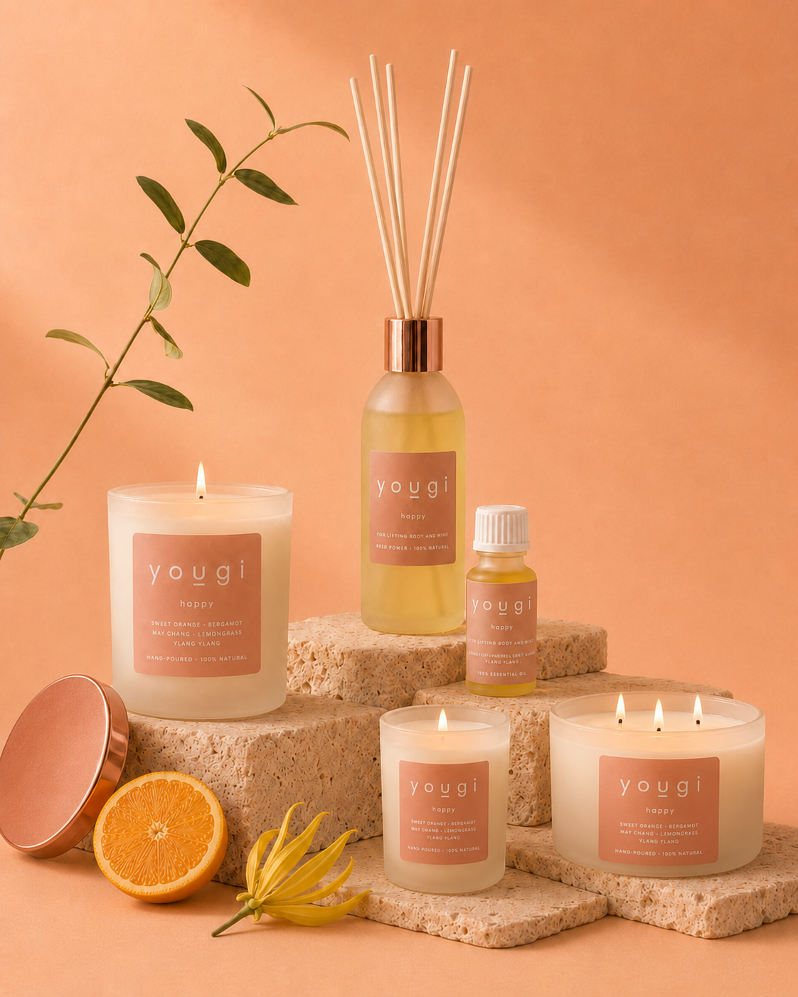 A coordinated set of Yougi home fragrance products arranged on stone blocks against a peach studio background, including a reed diffuser, candle, oil bottle, and two candles, with orange slices, a flower, and a leafy stem as styling props.