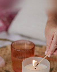 A frosted white glass candle being lit on a rattan tray, in the rose tones of the New Romantic blend