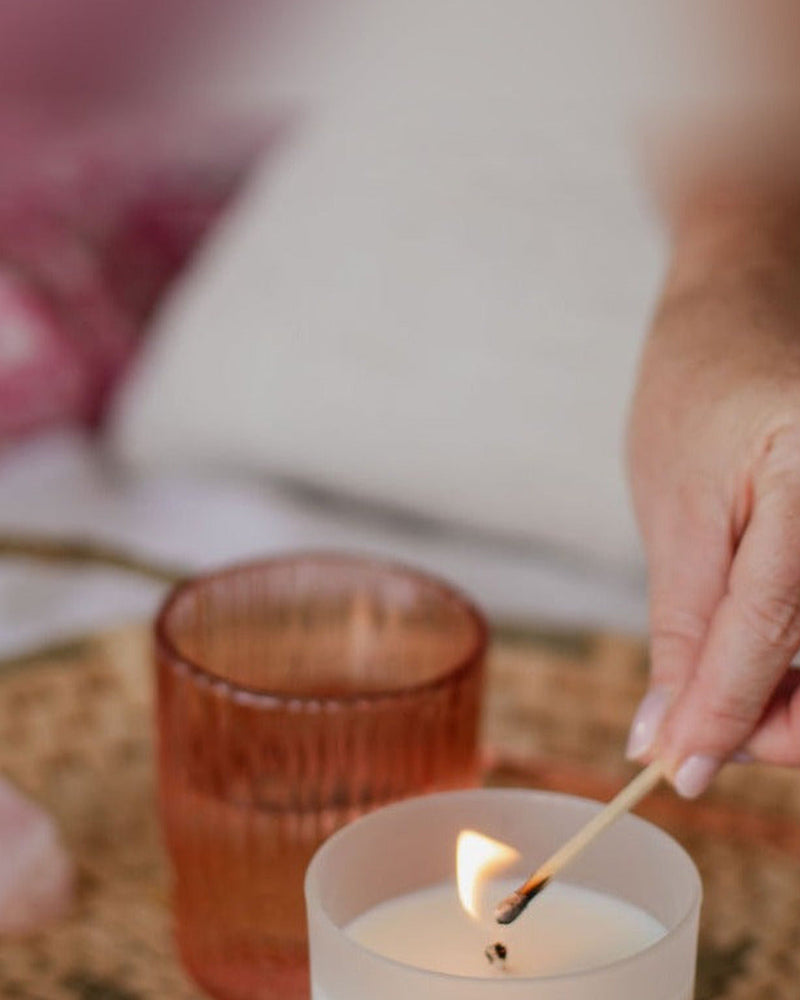 A frosted white glass candle being lit on a rattan tray, in the rose tones of the New Romantic blend