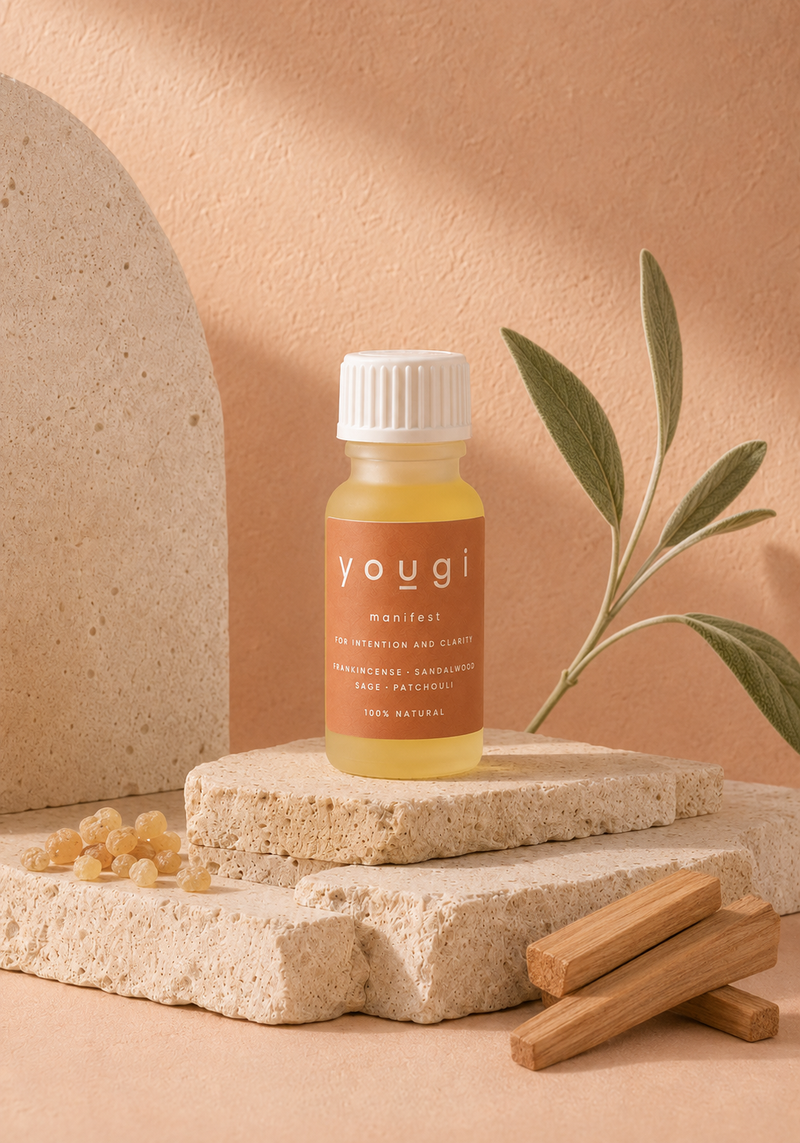 A small bottle of Yougi Manifest essential oil with a white cap and amber-brown label, styled on stacked stone blocks with a sage branch and wooden sticks against a warm peach background.
