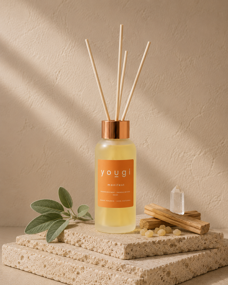 A Yougi reed diffuser bottle with a copper-toned cap and natural reeds, styled on stacked stone slabs with green leaves, wood pieces, and a crystal against a textured beige wall.