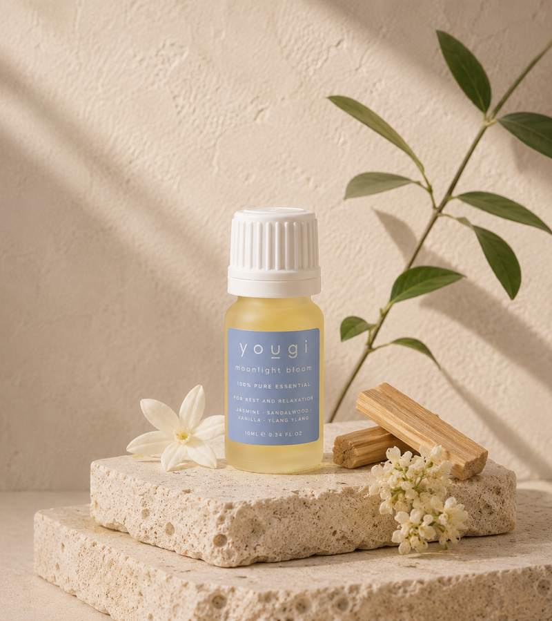 A small bottle of Youji Moonlight Cream essential oil with a white cap and light blue label, styled on a stone pedestal with a white flower, wood slices, and green leaves against a textured beige wall with soft shadows.