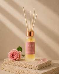 A reed diffuser bottle with pale yellow liquid, a pink label reading 'yougi', and four natural reed sticks, styled on stacked stone blocks with a pink rose and petals against a warm beige textured wall with soft diagonal shadows.