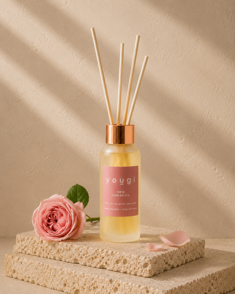 A reed diffuser bottle with pale yellow liquid, a pink label reading 'yougi', and four natural reed sticks, styled on stacked stone blocks with a pink rose and petals against a warm beige textured wall with soft diagonal shadows.