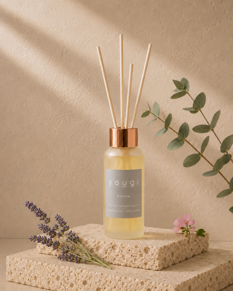 A reed diffuser bottle with a copper-toned cap and natural reed sticks, set on a textured stone block with dried lavender, a small pink flower, and eucalyptus leaves against a warm beige wall with soft shadows.