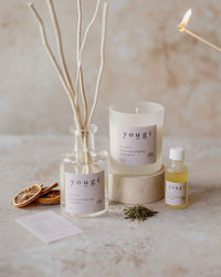 Happy candle being lit beside its reed diffuser and essential oil, with dried orange and lemongrass