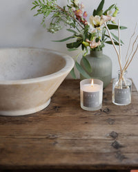 Soothe candle lit on a wooden washstand beside a stone basin and reed diffuser
